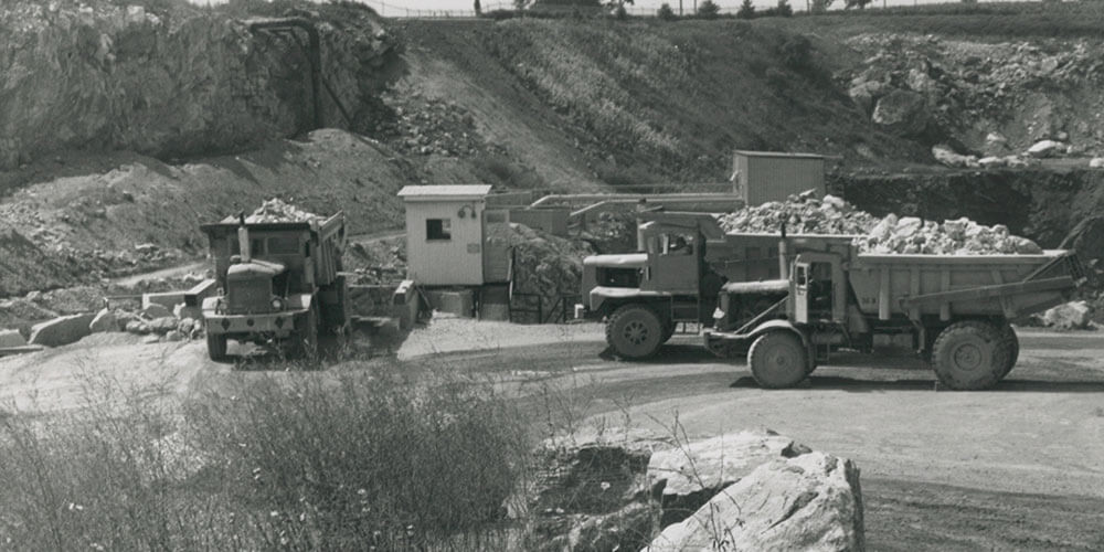 Black and white image of dump trucks at the bottom of the 2-tiered quarry in 1975